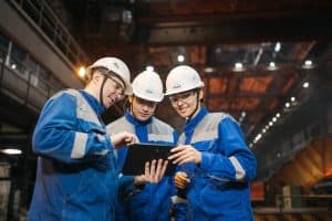 Tre arbetare i skyddshjälmar som diskuterar vid en industriell anläggning.