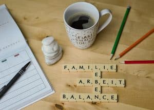 En kaffekopp, pennor och Scrabble-brickor som stavar ’FAMILIE, ARBEIT, BALANCE’ på ett bord.