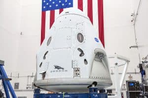 SpaceX Dragon-kapsel i en hangar med en amerikansk flagga i bakgrunden.