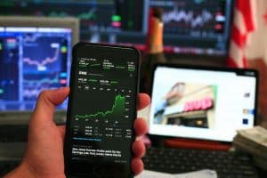 Hand som håller en smartphone som visar börsdata med datorskärmar i bakgrunden.