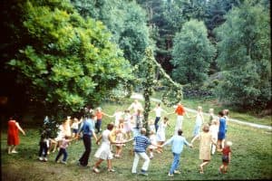 Människor som dansar runt en midsommarstång i en grön utomhusmiljö.