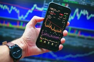 En hand håller en smartphone med aktiekurser på skärmen framför en datorskärm med grafer.